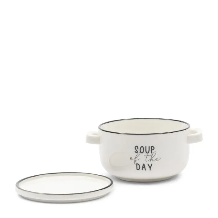 Podstawka na zupę w kolorze białym, ceramiczna, z dwoma uchwytami. Ma czarne brzegi oraz napis „SOUP OF THE DAY” na froncie. Przykrywa w kolorze białym leży obok. Produkt ustawiony na prostym tle.