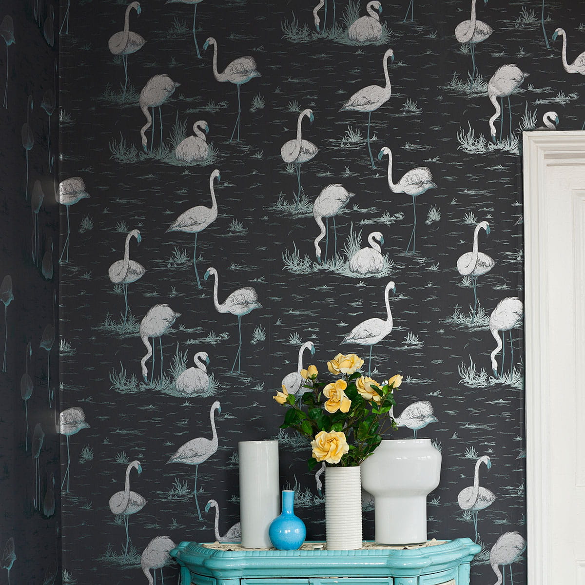 Tapeta Cole and Son Flamingos w kolorze białym i szarym w delikatny wzór flamingów na ciemnym tle. Produkt na ścianie, w tle widać niebieską komodę i wazon z żółtymi różami. Wzór ukazuje flamingi stojące i siedzące w trawie.