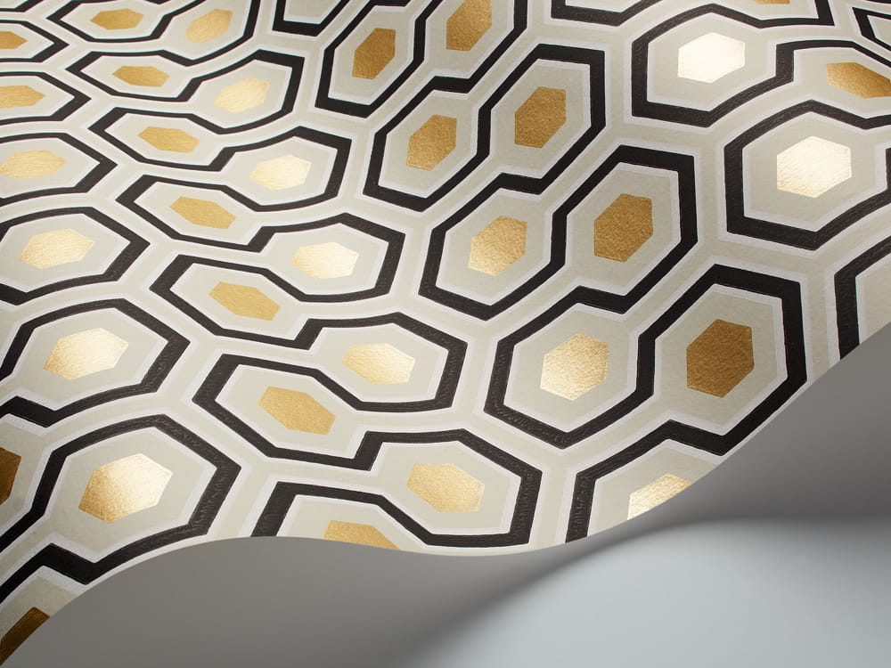 Tapeta Cole and Son Hicks' Hexagon w kolorze miękkiej oliwki, złota i węgla. Wzór z sześciokątami i drobnymi detalami w złotym wykończeniu. Produkt leży na gładkiej powierzchni, z częściowo zwiniętym brzegiem, ukazując elegancki design.