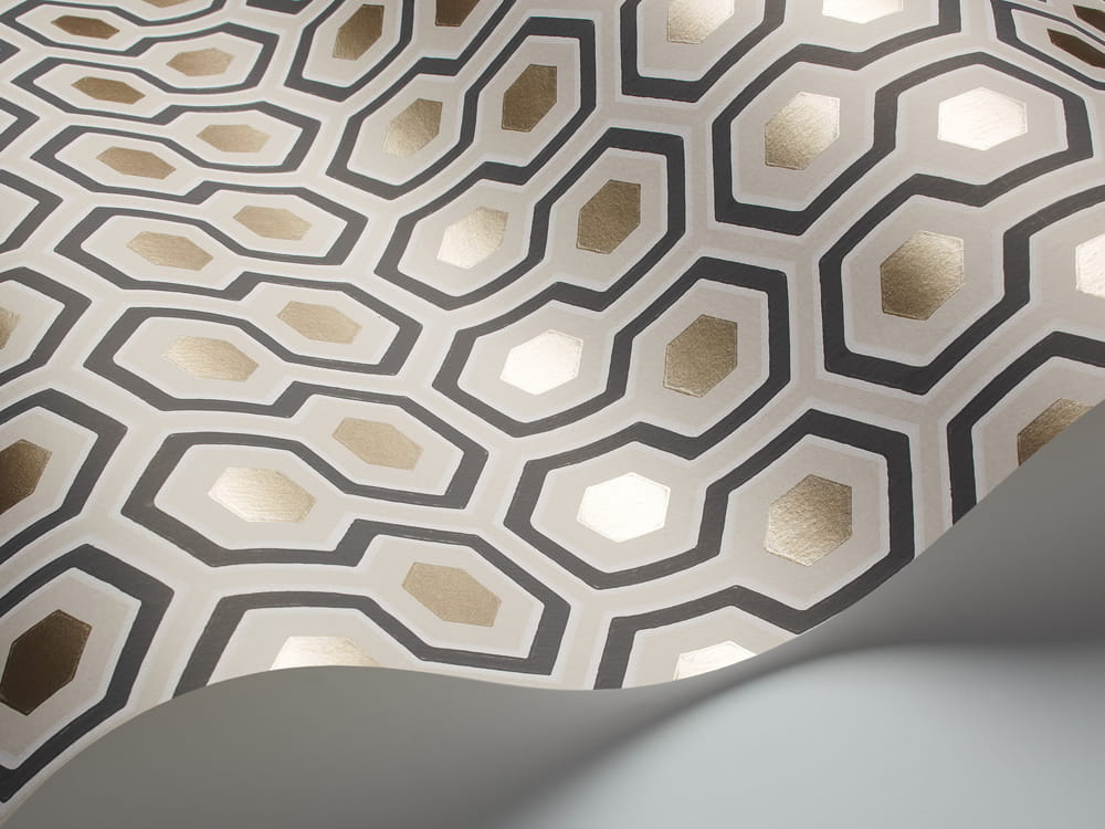 Tapeta Cole and Son Hicks' Hexagon w odcieniach beżu, złota i antracytu, z geometrycznym wzorem heksagonalnym. Widoczna struktura i błyszczące elementy w złotym kolorze na matowym tle. Produkt lekko zwinięty, ukazujący detal wzoru.