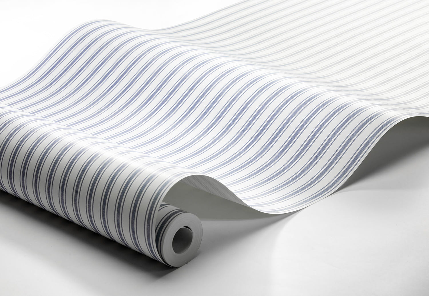 Tapeta Marstrand II Aspo Stripe w kolorze błękitno-białym, z delikatnym, falistym wzorem pasków. Produkt leży na gładkim, jasnym tle, zwinięty na jednym końcu, co ukazuje jego fakturę i design.