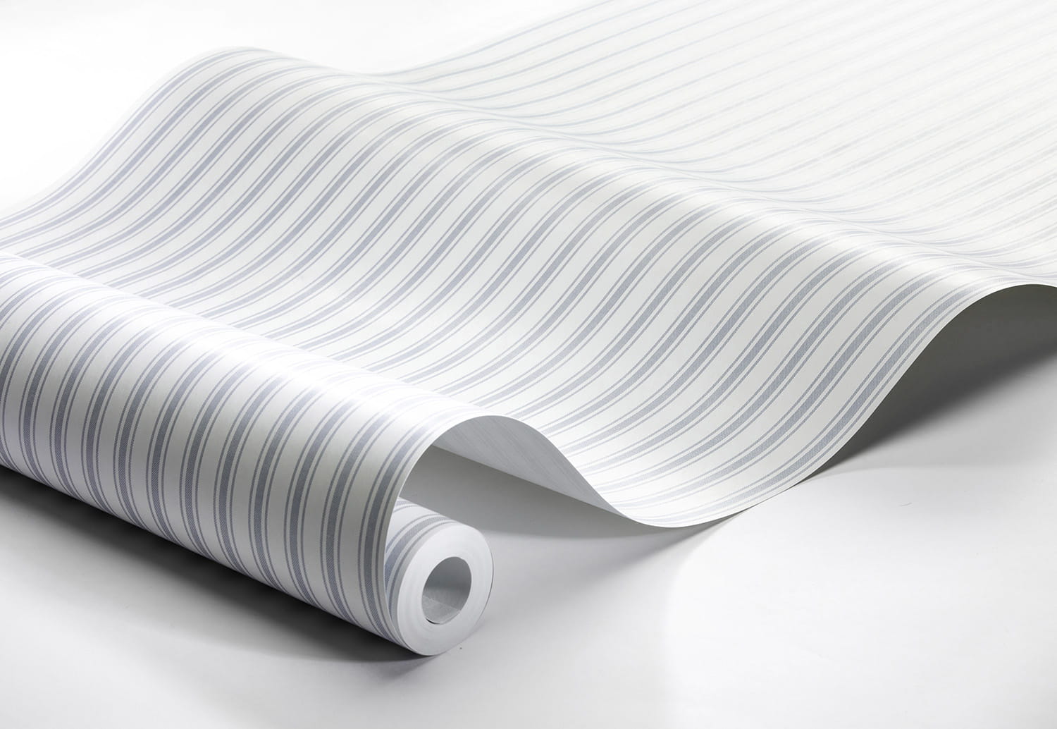 Tapeta Marstrand II Aspo Stripe w kolorze jasnoniebieskim i białym, z falistym wzorem w paski. Produkt leży na stole, z rolką widoczną z jednej strony, ukazując teksturę i wzór. Tło jest jasne, co podkreśla kolorystykę tapety.