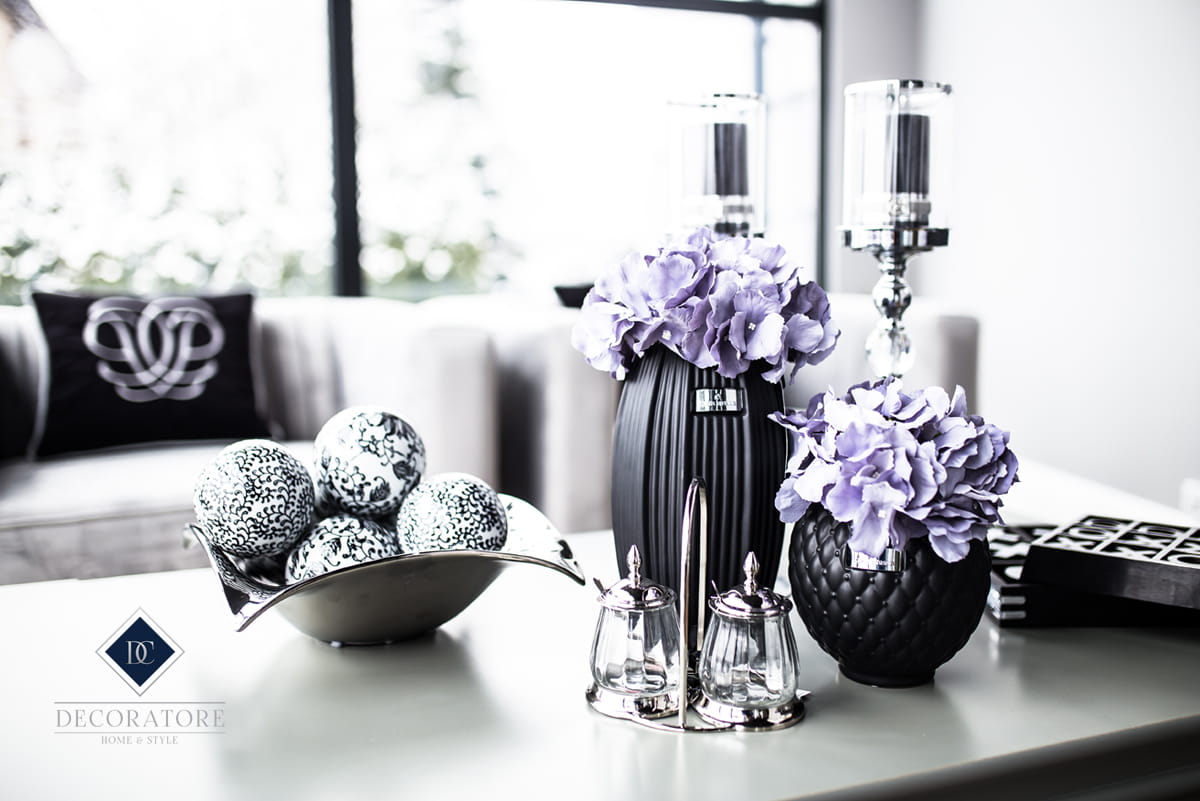 Czarna świeca Lene Bjerre umieszczona w eleganckim wazonie z fioletowymi kwiatami. Produkt na stole, w otoczeniu dekoracyjnych kul w metalowej misce oraz szklanych pojemników na przyprawy. Tło jasne, minimalistyczne.