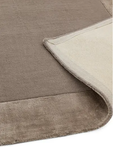 Dywan Ascot w kolorze taupe, z widoczną miękką teksturą i obrzeżem. Zrolowany brzeg ukazuje gładką spód. Produkt oferowany w różnych rozmiarach, idealny do aranżacji wnętrz.