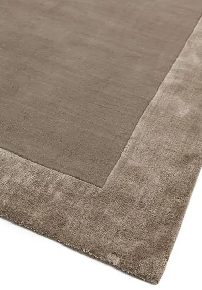 Dywan Ascot w kolorze taupe, z gładką powierzchnią i miękką, teksturowaną ramką. Widoczny detal brzegów, bez wzorów. Produkt leżący na podłożu, dostępny w różnych rozmiarach.