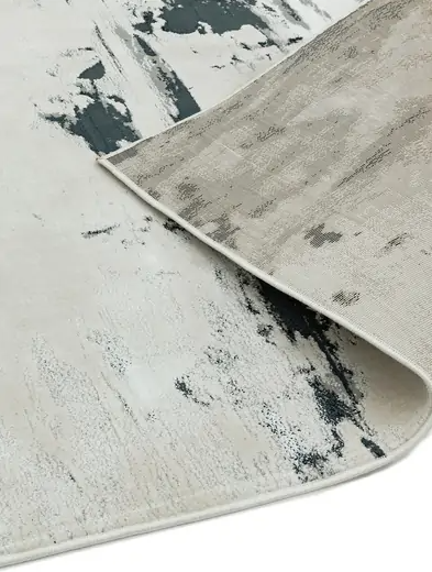 Dywan Aurora Glacier w odcieniach beżu i szarości z abstrakcyjnym wzorem. Widoczny rąbek, materiał o teksturowanej strukturze. Produkt leży na powierzchni, lekko podwinięty w jednym rogu. Dostępny w różnych rozmiarach.