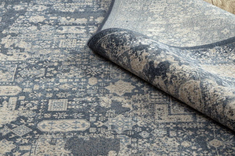 Dywan Nuet II w kolorze granatowym i beżowym, złożony w rogu, ukazuje wzór w stylu vintage z geometrycznymi i florystycznymi motywami. Wykonany z materiału o gładkiej fakturze, dostępny w różnych rozmiarach.