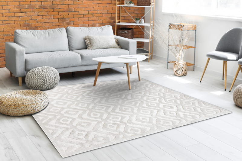 Dywan Santi Sznurkowy w kolorze beżowym, z wzorem zygzaków, leży na jasnej podłodze. Widoczne są miękkie pufy oraz szary, tapicerowany sofa. Produkt ukazany z przodu w aranżacji wnętrza z ceglaną ścianą, tworząc przytulną przestrzeń.