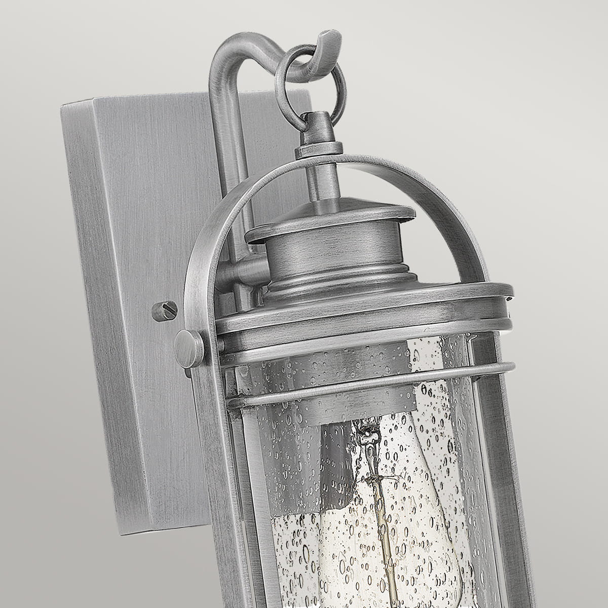 Kinkiet zewnętrzny Benev S w kolorze aluminium, o wymiarach 16x16x29 cm, zawieszony na ścianie. Podświetlenie przez szklany klosz z bąbelkowymi detalami, a także metalowa konstrukcja z dekoracyjnym uchwytem. Produkt na jednolitym tle.