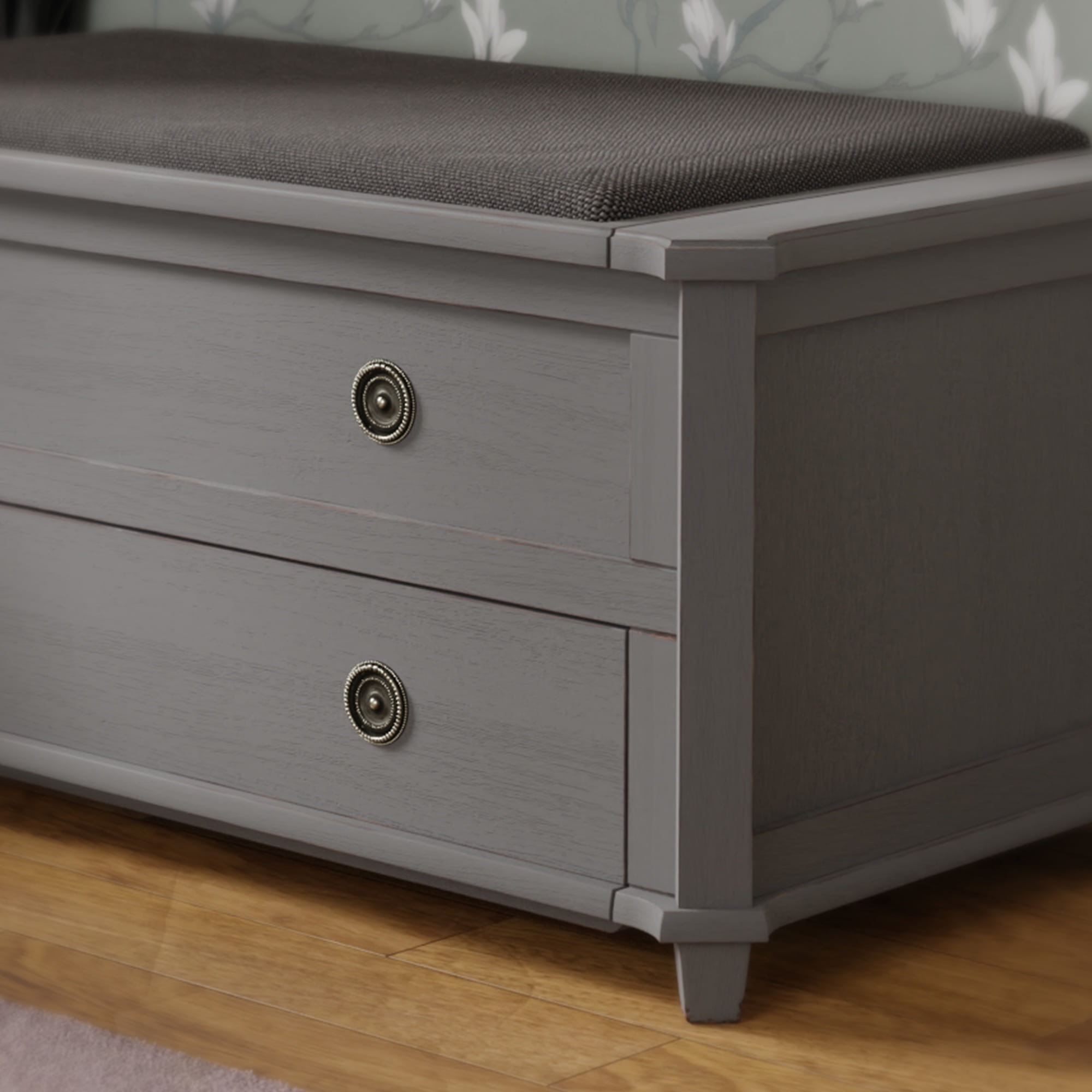 Szafka drewniana na buty Henshaw Laura Ashley w kolorze szarym z dwoma szufladami. Widoczna ciemnoszara poduszka na górze, okrągłe uchwyty w kolorze złotym. Produkt stoi na drewnianej podłodze w jasnym otoczeniu.