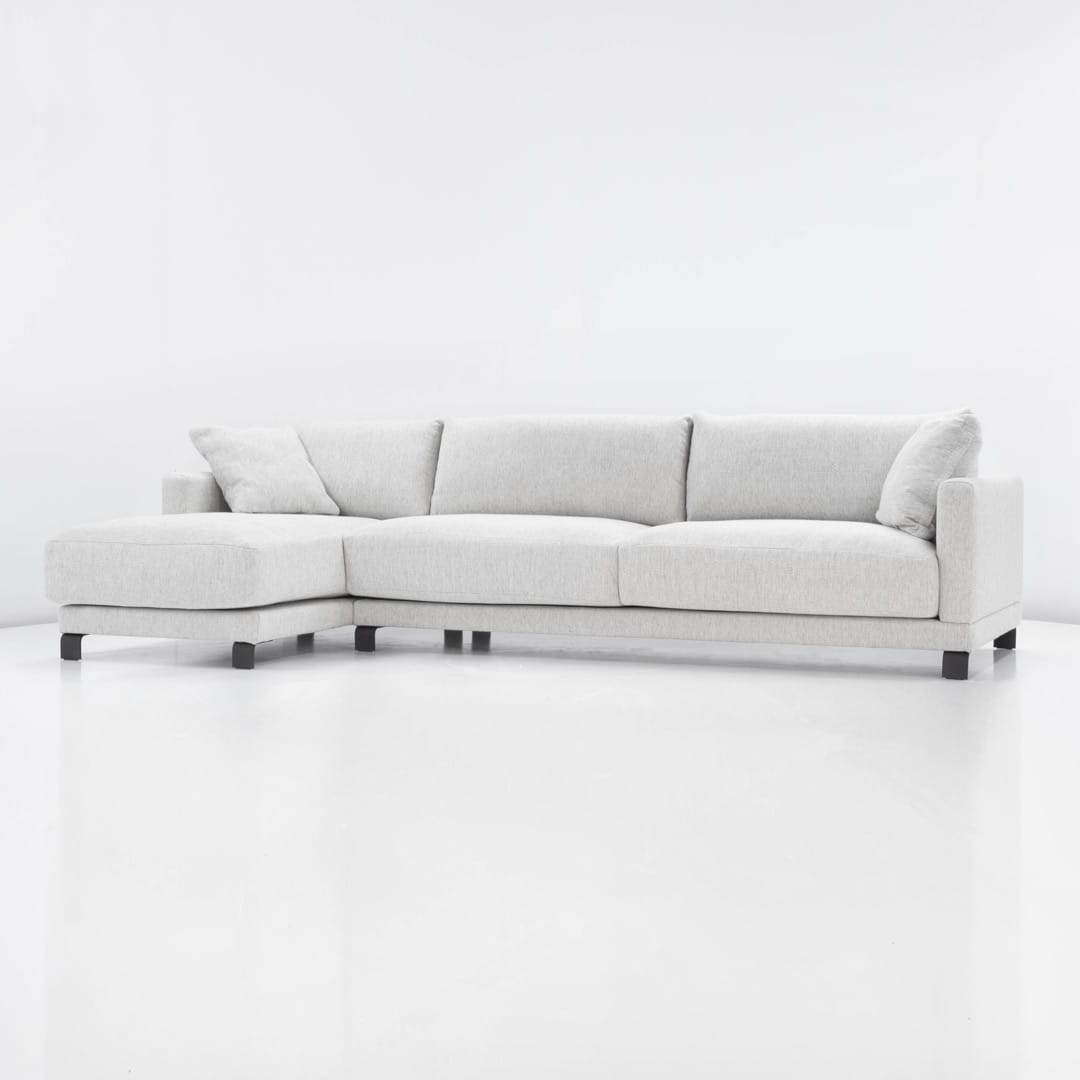 Komfortowa sofa narożna w jasnoszarym kolorze, z dwiema poduchami, o wymiarach 260x160 cm i wysokości 85 cm. Ujęcie z boku, produkt ułożony poziomo na białym tle, prezentujący minimalistyczny design i funkcjonalne kształty.