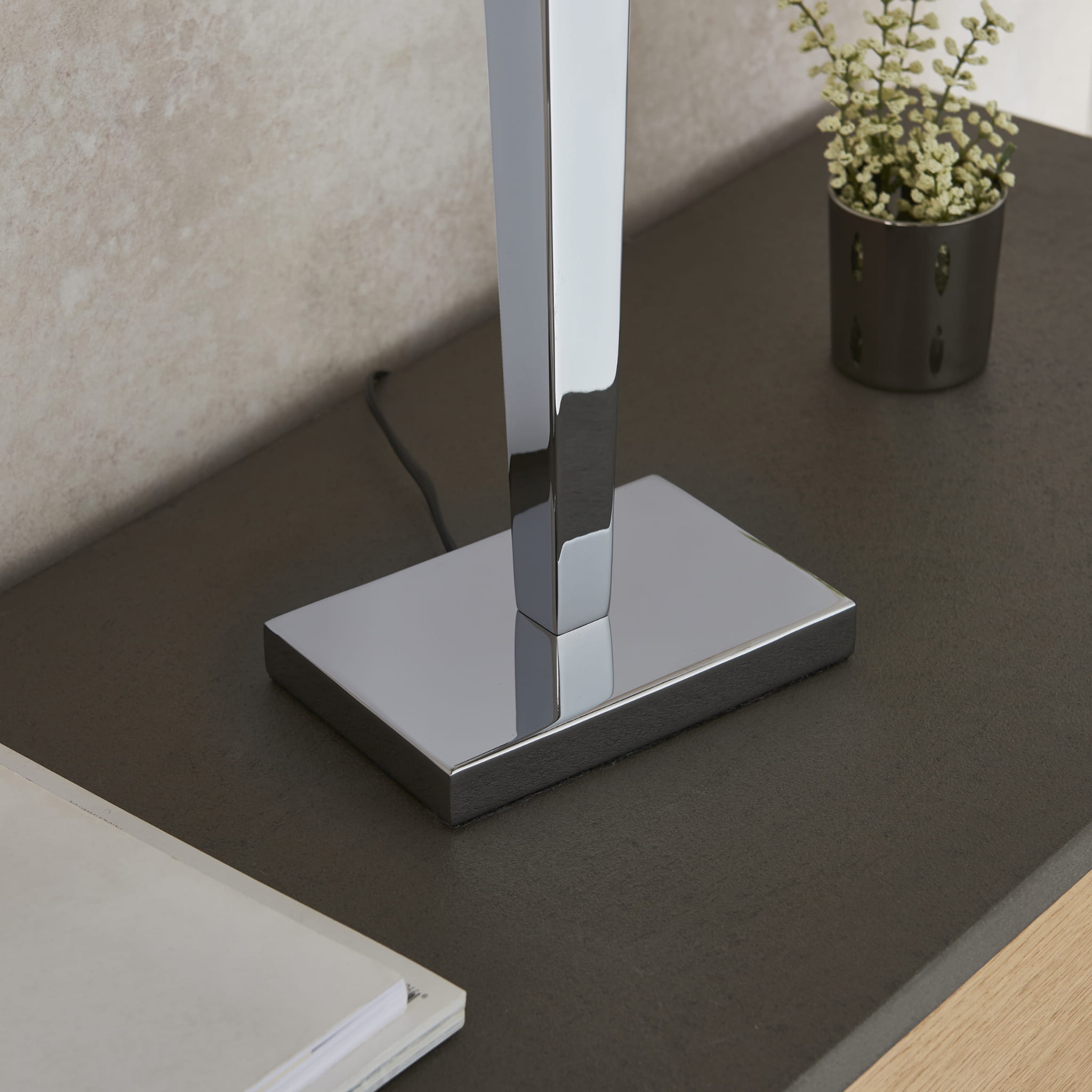 Lampa stołowa Moreto w kolorze chromu, z prostą, metalową podstawą w kształcie kwadratu. Górna część lampy jest smukła i elegancka. Produkt umieszczony na ciemnym, matowym blacie, obok leży notes oraz mały kwiatek w doniczce.