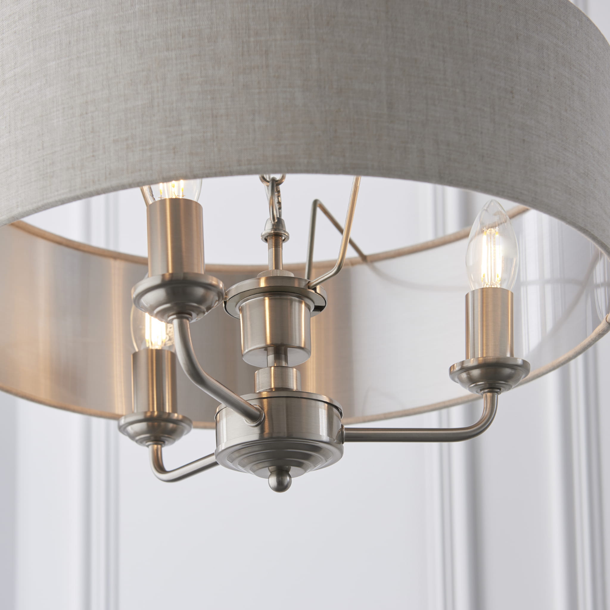 Lampa sufitowa wisząca Highclere o wysokości 142 cm. Metalowe elementy w chromowanym wykończeniu oraz tekstylny klosz w jasno-szarym kolorze. Produkt wiszący, z trzema żarówkami widocznymi na zdjęciu. Tło proste, białe.