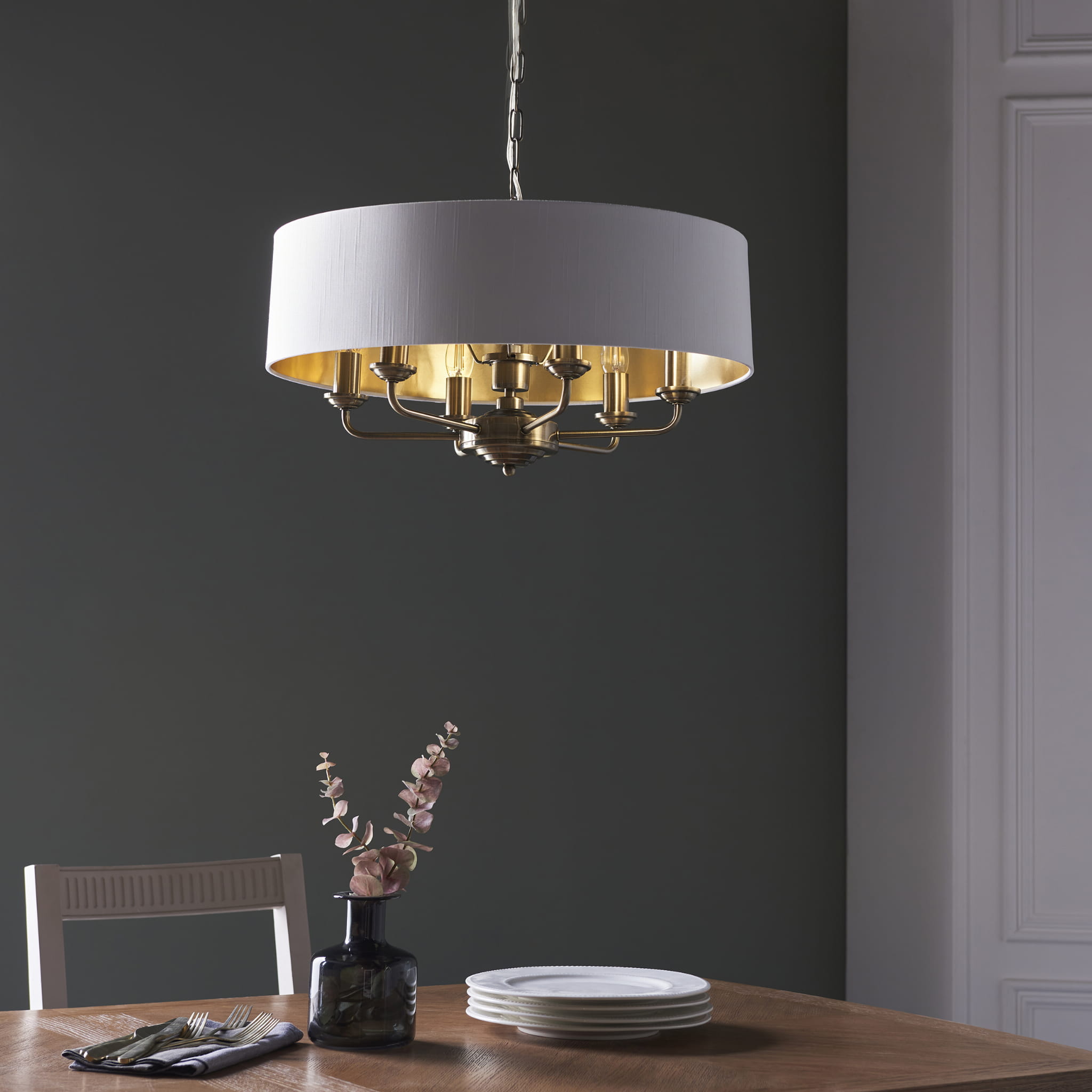 Lampa sufitowa wisząca Highclere wykonana z mosiądzu, z tkaninowym abażurem w kolorze białym od spodu ze złotym wykończeniem. Produkt wisi nad drewnianym stołem, w tle ciemna ściana. Widoczne dwa kształty talerzy oraz kwiat w wazonie na stole.