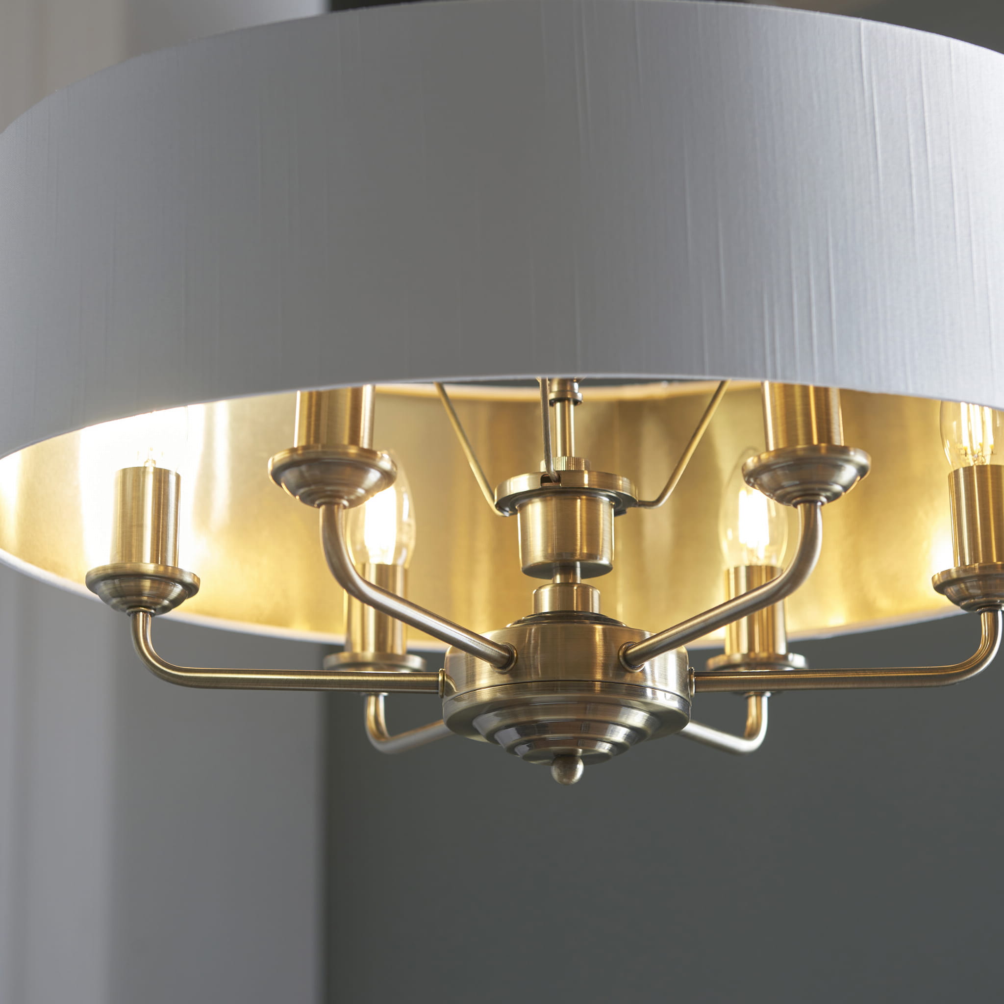 Lampa sufitowa wisząca Highclere w kolorze mosiądzu z tekstylnym abażurem. Produkt z sześcioma ramionami, umieszczony w centralnym punkcie. Abażur ma kolor jasnoszary, a wnętrze metaliczne, co podkreśla elegancję lampy. Kąt ujęcia: z góry.
