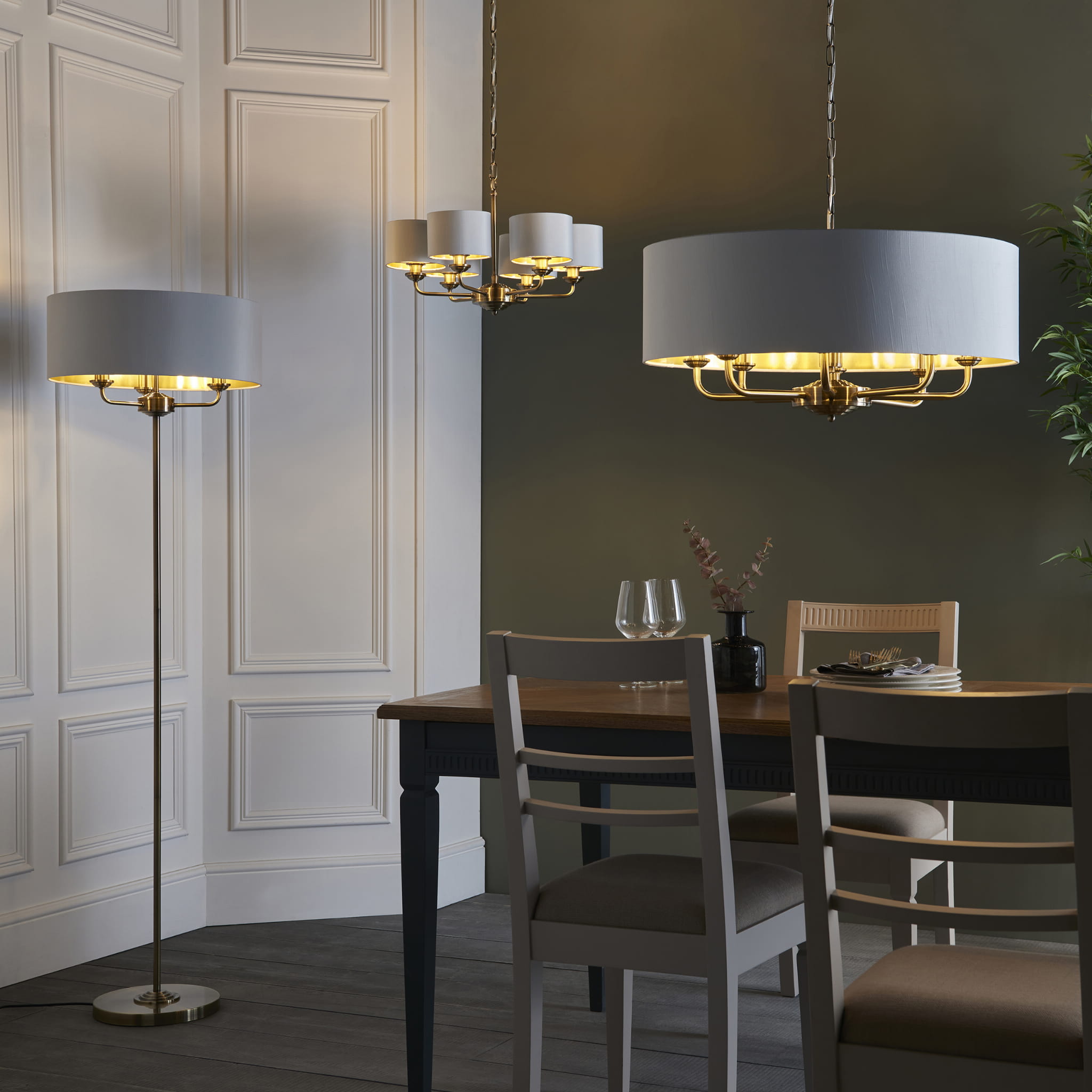 Lampa podłogowa Highclere w kolorze mosiądzu, wysokość 160 cm, umieszczona w aranżacji wnętrza. Produkt stojący z okrągłym kloszem w jasnym odcieniu. W tle widoczny stół z krzesłami oraz lampy sufitowe, co podkreśla kontekst użytkowania.