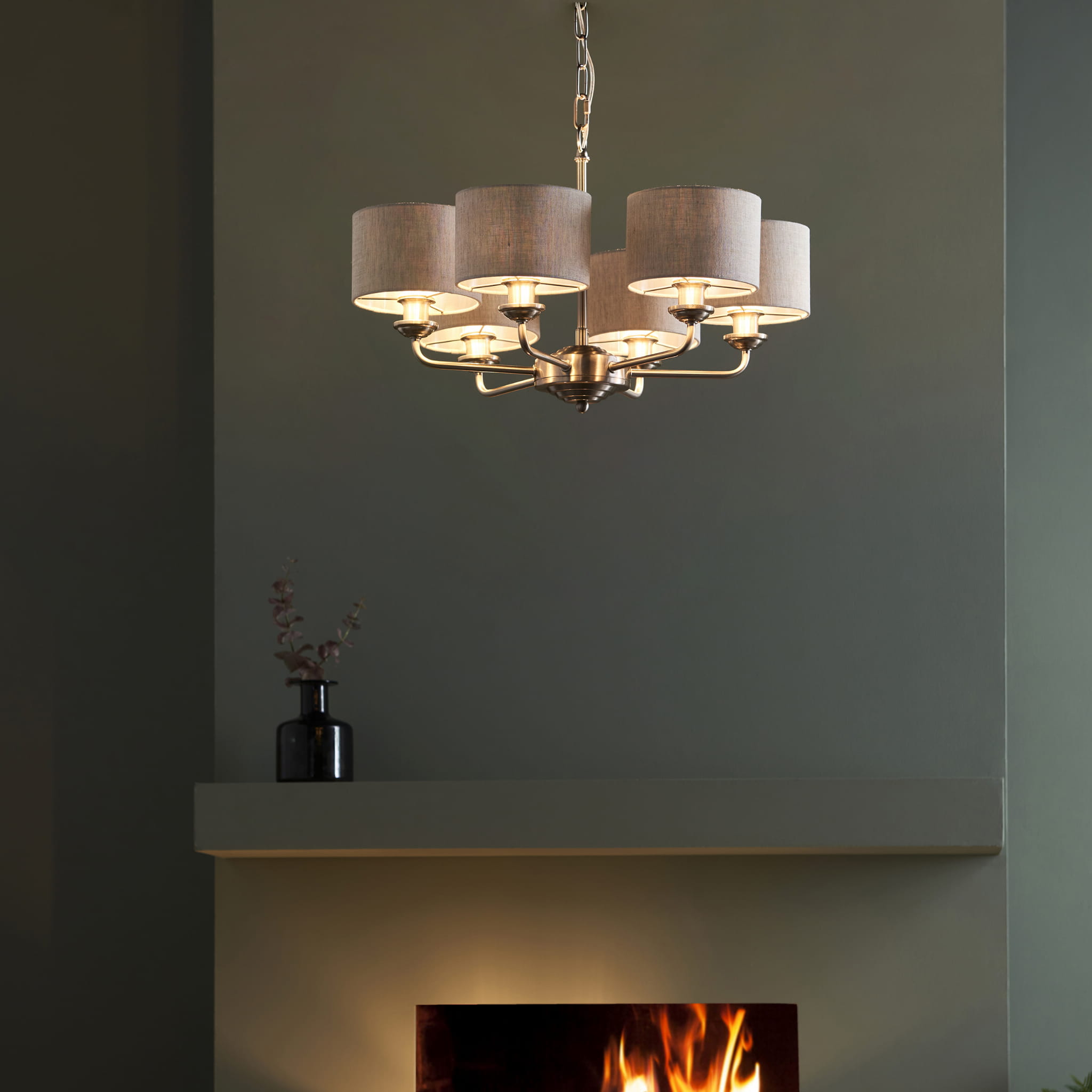 Lampa sufitowa wisząca Highclere w kolorze chromu i jasnego lnu, z sześcioma kloszami w formie cylindrów. Produkt wiszący, z metalową konstrukcją i oświetleniem LED. Umieszczony w kolorowej aranżacji wnętrza z kominkiem w tle.