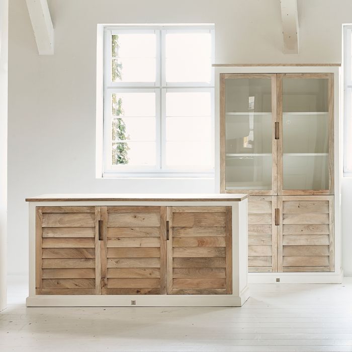 Komoda Pacifica Riviera Maison w kolorze naturalnego drewna z jasnymi, pionowymi panelami. Produkt stoi w prostokątnej formie, z trzema drzwiami i metalowymi uchwytami. Tło jasne, z oknem w tle. Wymiary: 182x50x91 cm.