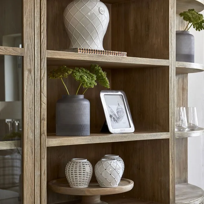 Ceramiczna waza amfora w kolorze białym, z gładką powierzchnią i zdobieniem w formie rombów, wysoka na 27 cm, umieszczona na drewnianej półce obok szarych wazonów. Ujęcie frontowe, produktu w otoczeniu dekoracji i ramki ze zdjęciem.