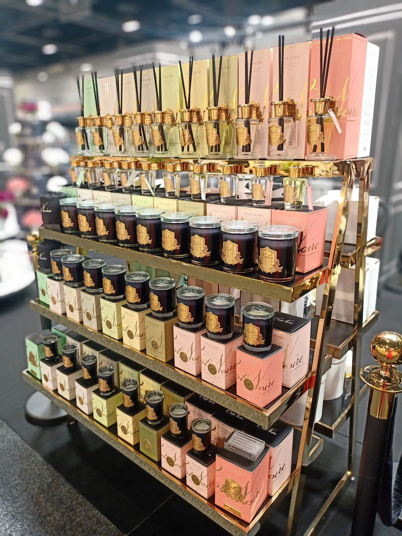 Stojąca półka z zapachowymi świecami marki Cote Noire. Na górze umieszczone są szklane flakony z czarnymi rurkami, poniżej elegantne, okrągłe słoiki w kolorach czarnym, zielonym, różowym i złotym. Produkt w zestawieniu na jednolitym tle.