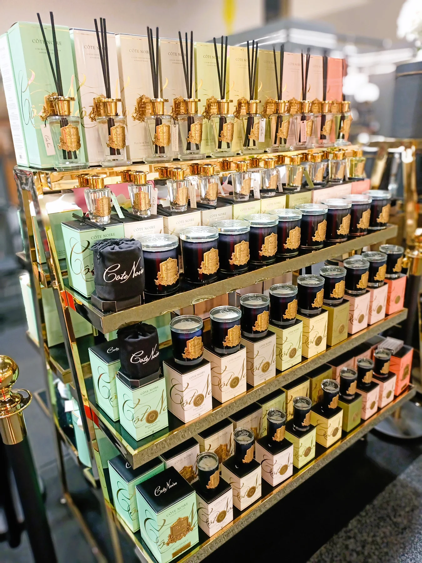 Sojowa świeca zapachowa Blonde Vanilla 185g umieszczona na złotej półce, w otoczeniu innych świec i dyfuzorów. Świeca ma czarny słoik z złotą dekoracją. W tle widoczne pastelowe opakowania produktów Côté Noire w różnych kolorach.