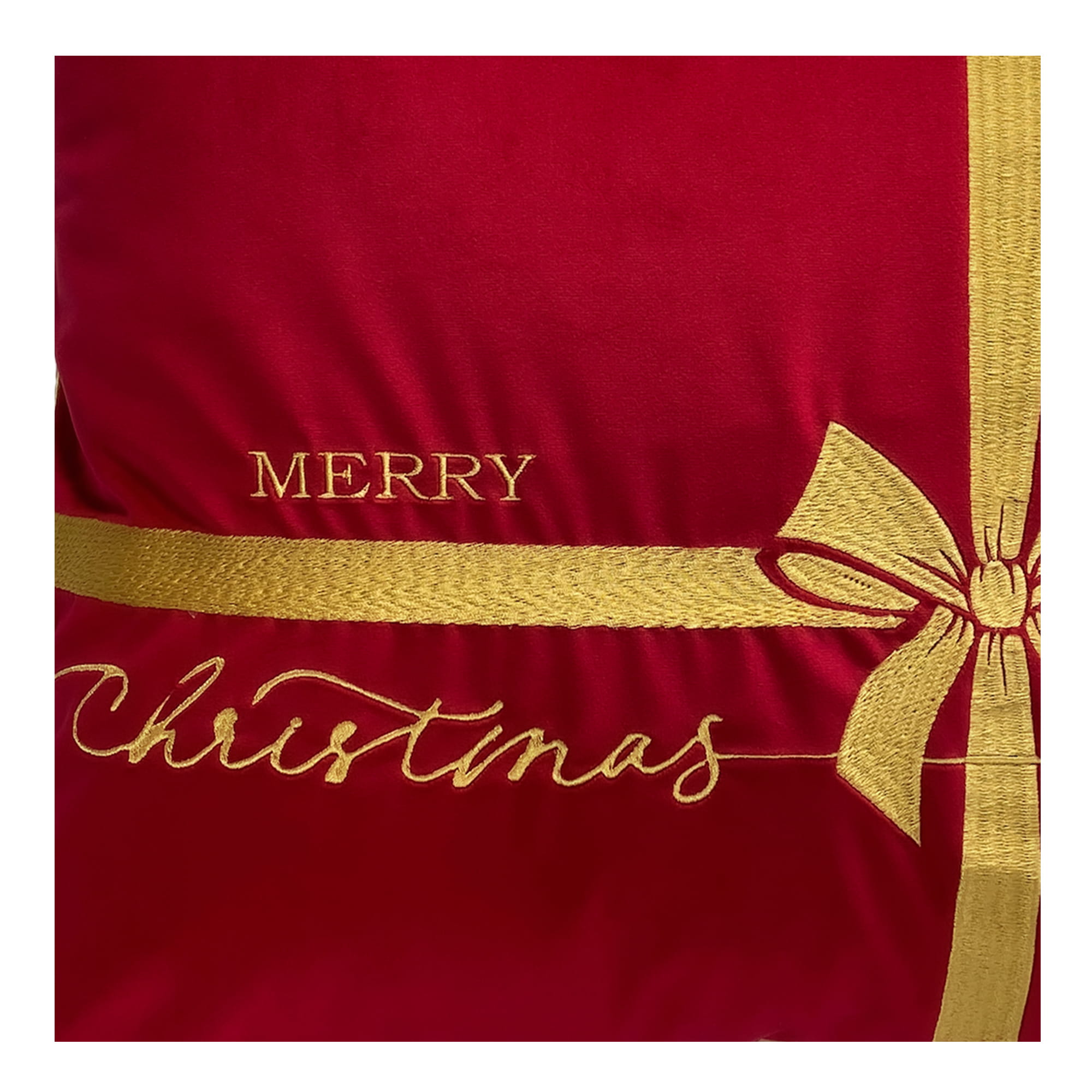 Poduszka dekoracyjna w czerwonym kolorze z haftem w złotym kolorze, zawierająca napis MERRY CHRISTMAS. Produkt leżący, zbliżenie na detal haftu, prezentujący ozdobną kokardę i sztuczny materiał. Idealna do dekoracji w okresie świątecznym.