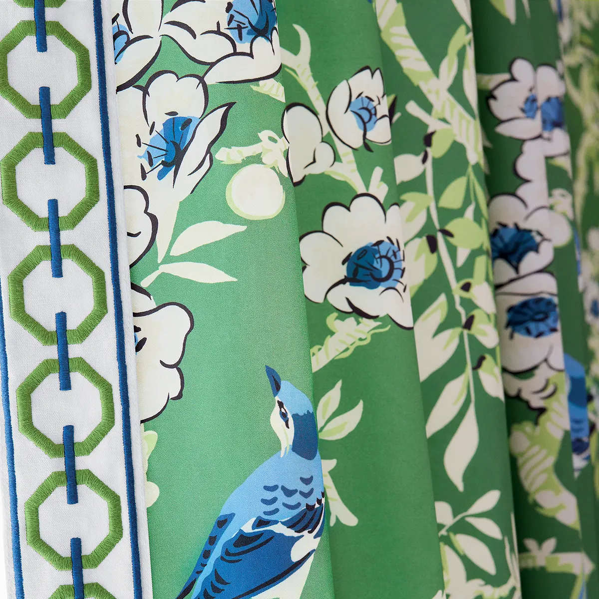Tapeta Thibaut Anna French YUKIO w odcieniach zieleni z dużymi białymi kwiatami i niebieskim ptakiem. Widoczny pasek z geometrycznym wzorem w kolorze zielonym i niebieskim. Ujęcie z przodu, produkt stoi w pozycji rozłożonej.