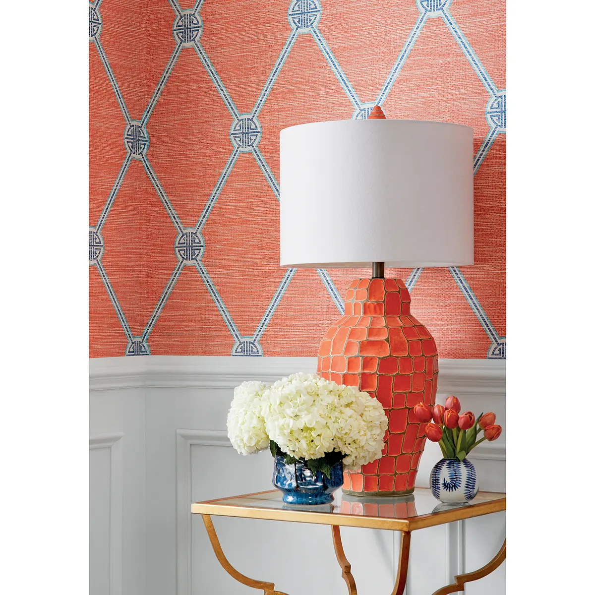 Tapeta Thibaut Anna French TURNBERRY TRELLIS w kolorze czarnym i beżowym z geometrycznym wzorem w formie rombów. Zasłonięta lampa na stoliku ze szklanym blatem, obok kwiaty w wazonie. Ujęcie frontalne, produkt umieszczony w eleganckim wnętrzu.