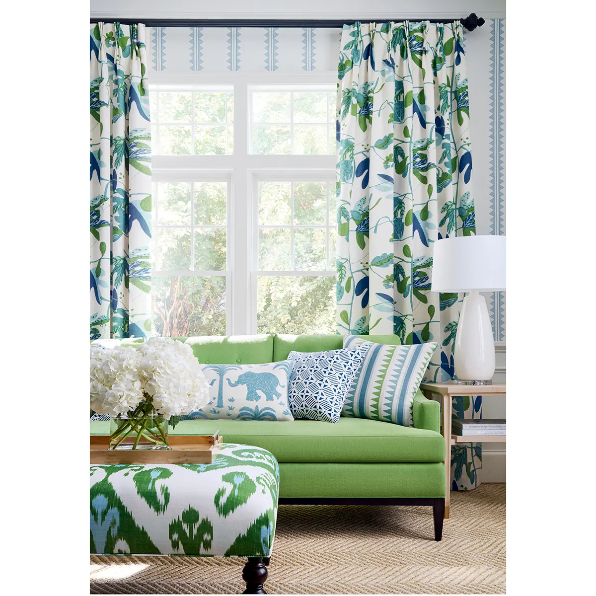 Tapeta Thibaut Anna French MATISSE LEAF w kolorach żółtym, niebieskim i pomarańczowym, zdobiona motywem liści. Produkt w formie zwisających zasłon, ukazany z boku w jasnym wnętrzu z zieloną kanapą i poduszkami, w harmonii z dekoracjami.