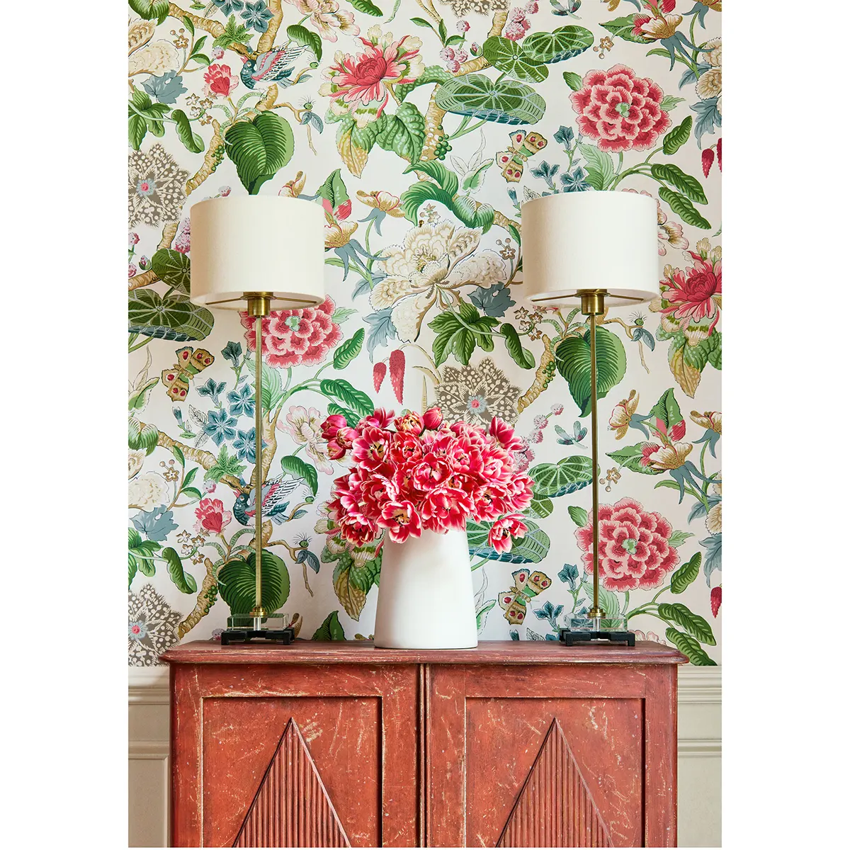 Tapeta Thibaut Anna French HILL GARDEN w jasno-niebieskim kolorze, wzór florystyczny z różowymi i zielonymi akcentami. Produkt na tle z lampami i wazonem z kwiatami, umieszczony na czerwonym meblu z detalami w geometrycznym stylu.