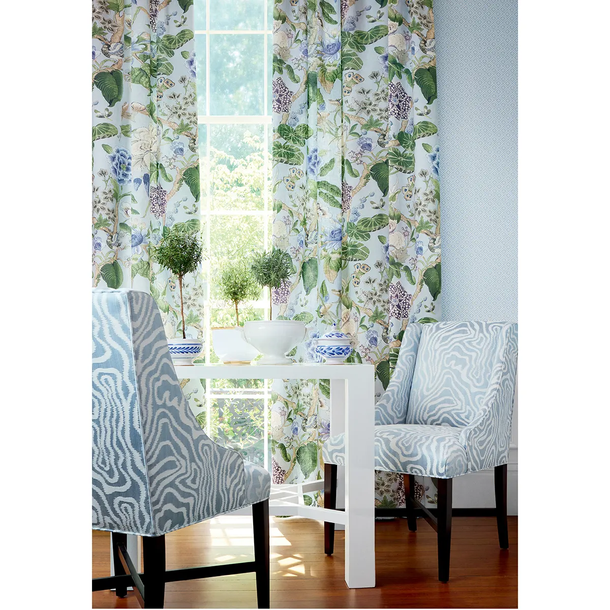 Tapeta Thibaut Anna French HOLIDAY TRELLIS w błękitnym kolorze, z delikatnym wzorem w kwiaty. Widok z przodu; produkt umieszczony na tle jasnej ściany oraz okna. Używana w aranżacji z białym stołem, niebieską misą i dekoracyjnymi roślinami.