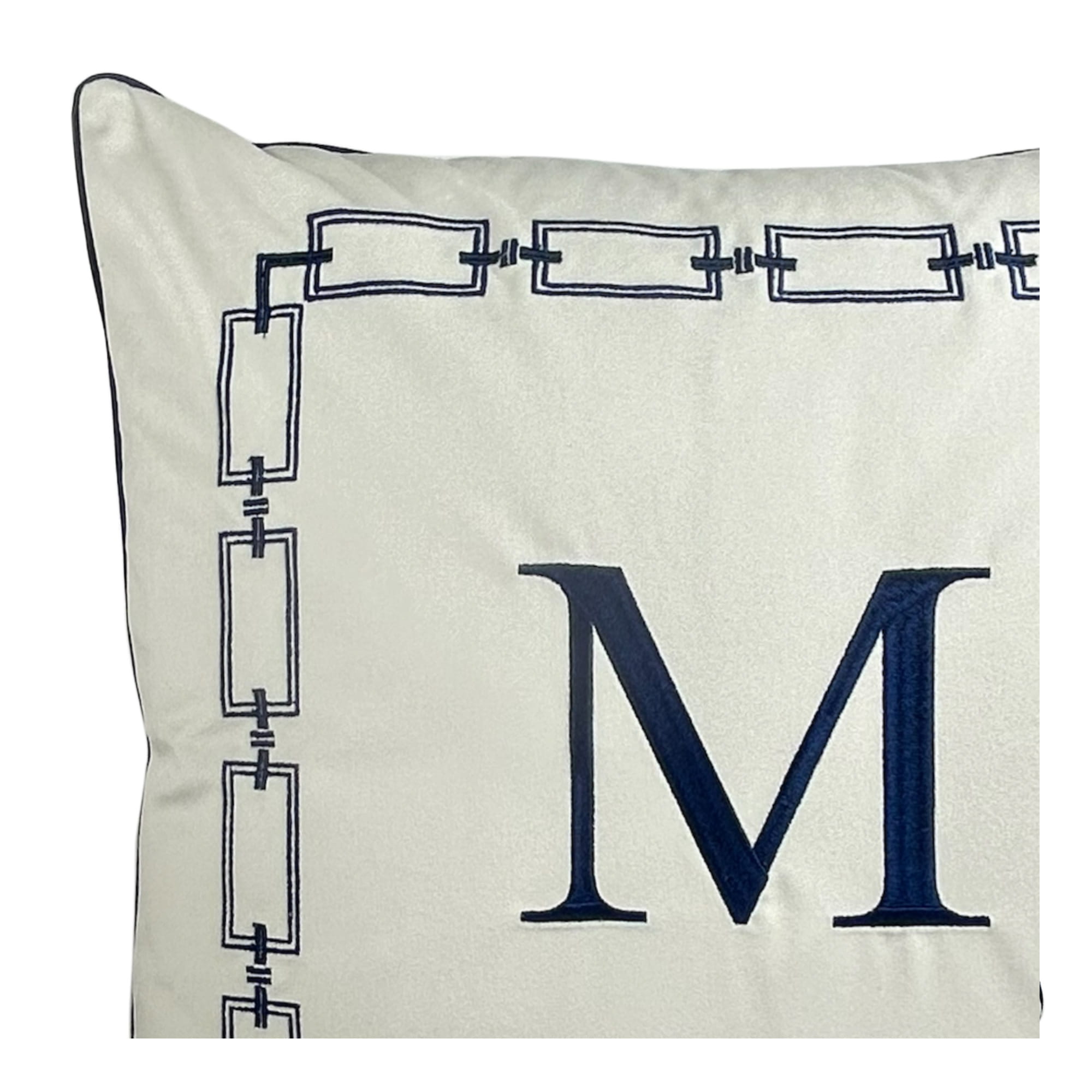 Poduszka dekoracyjna w kolorze jasnym z granatowym haftem monogramu M. Wokół monogramu znajduje się stylowy, ozdobny wzór ramkowy. Produkt leży płasko na białym tle, ukazując detale haftu i fakturę tkaniny.