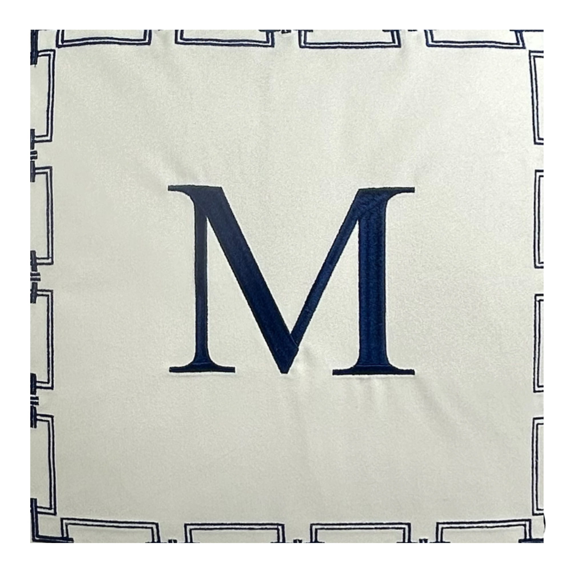 Poduszka haftowana dekoracyjna monogram w kolorze podstawowym białym z granatowym monogramem M pośrodku. Produkt leży płasko, nałożony na lekko teksturowane tło. Widoczny jest także granatowy haftujący wzór w formie prostokątów.