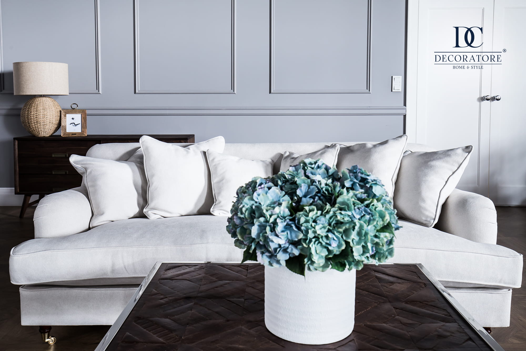 Trzyosobowa sofa Brighton w kolorze jasnobeżowym z poduszkami w kształcie prostokąta. Widok frontalny, produkt rozłożony. W tle widoczna szara ściana oraz stolik z kwiatami. Części drewniane w odcieniach ciemnego brązu.