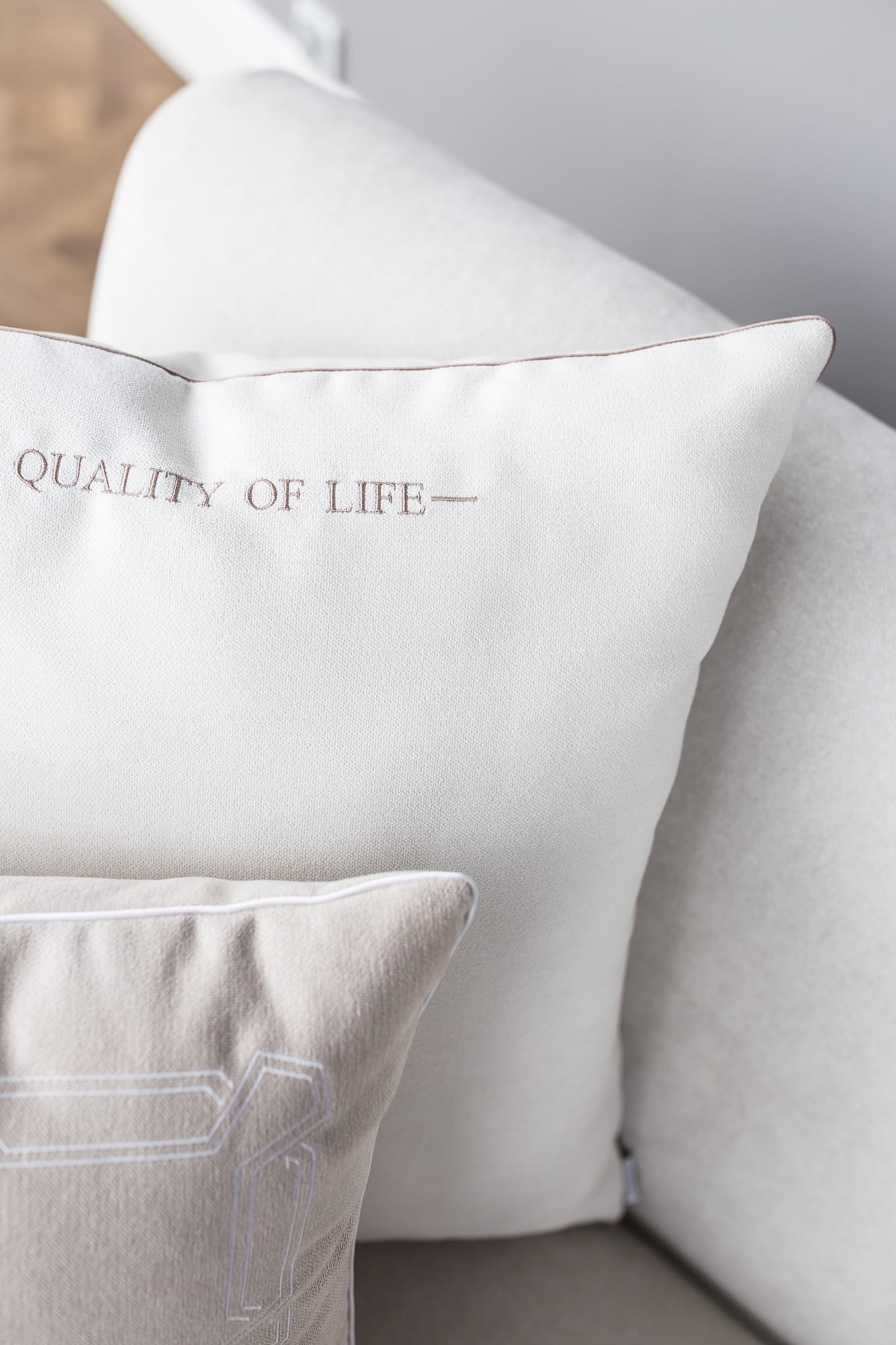 Poduszka dekoracyjna w jasnym odcieniu z napisem QUALITY OF LIFE w kolorze taupe, na tle beżowej kanapy. Z przodu widoczna jest także mniejsza poduszka w odcieniu szaro-beżowym z delikatnym wzorem. Produkty leżą na kanapie, z bliska.