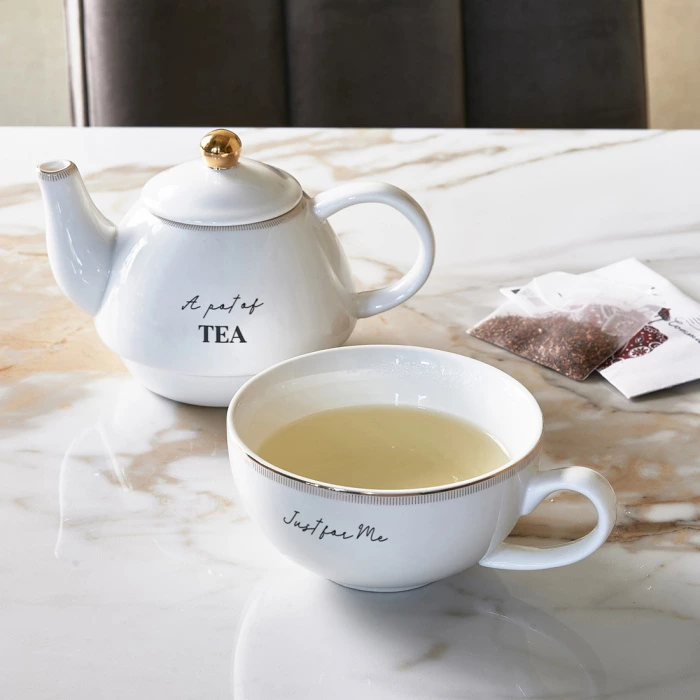 Czajniczek do herbaty w kolorze białym z złotym uchwytem, z widocznym napisem „A pot of TEA”. Obok filiżanka z napisem „Just for Me”, wypełniona herbatą. Tło marmurowe, produkt stoi na stole. W kadrze widoczne torebki herbaty.