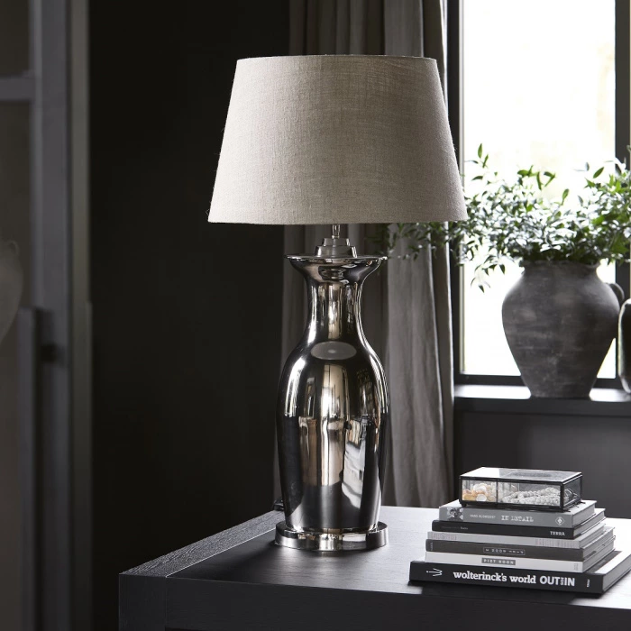 Podstawa lampy w kształcie stożka, wykonana z błyszczącego metalu w kolorze srebrnym. Użyto abażura z beżowego materiału. Produkt stoi na czarnej powierzchni, w tle widoczne rośliny oraz książki, tworzące dekoracyjną aranżację.