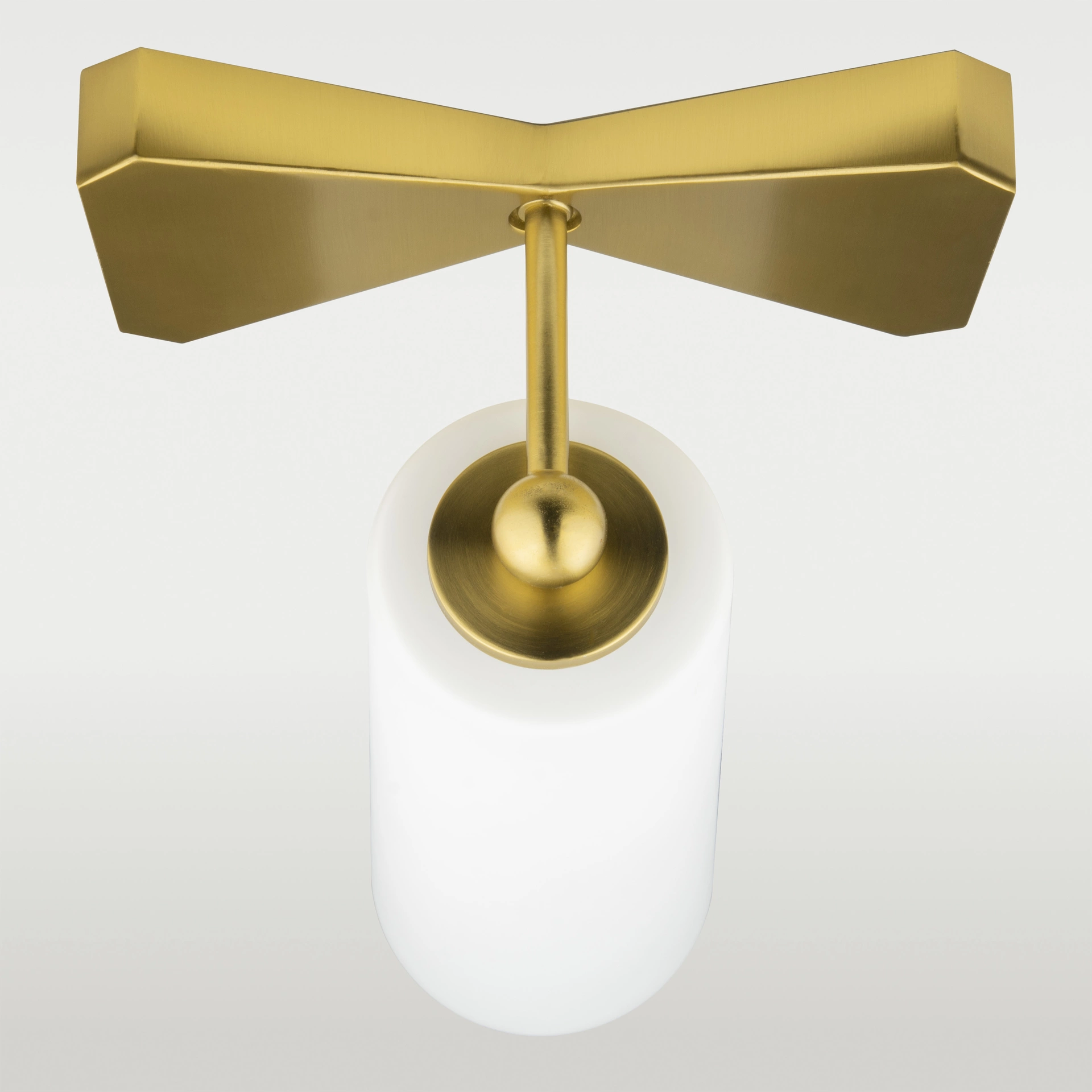 Kinkiet Bow w kolorze mosiądzu, z cylindrycznym kloszem z białego szkła. Ujęcie z góry, produkt wiszący, ukazujący detale konstrukcji mocującej i eleganckie wykończenia. Jasne tło podkreśla minimalistyczny design lampy. Wymiary: 14x18x32 cm.