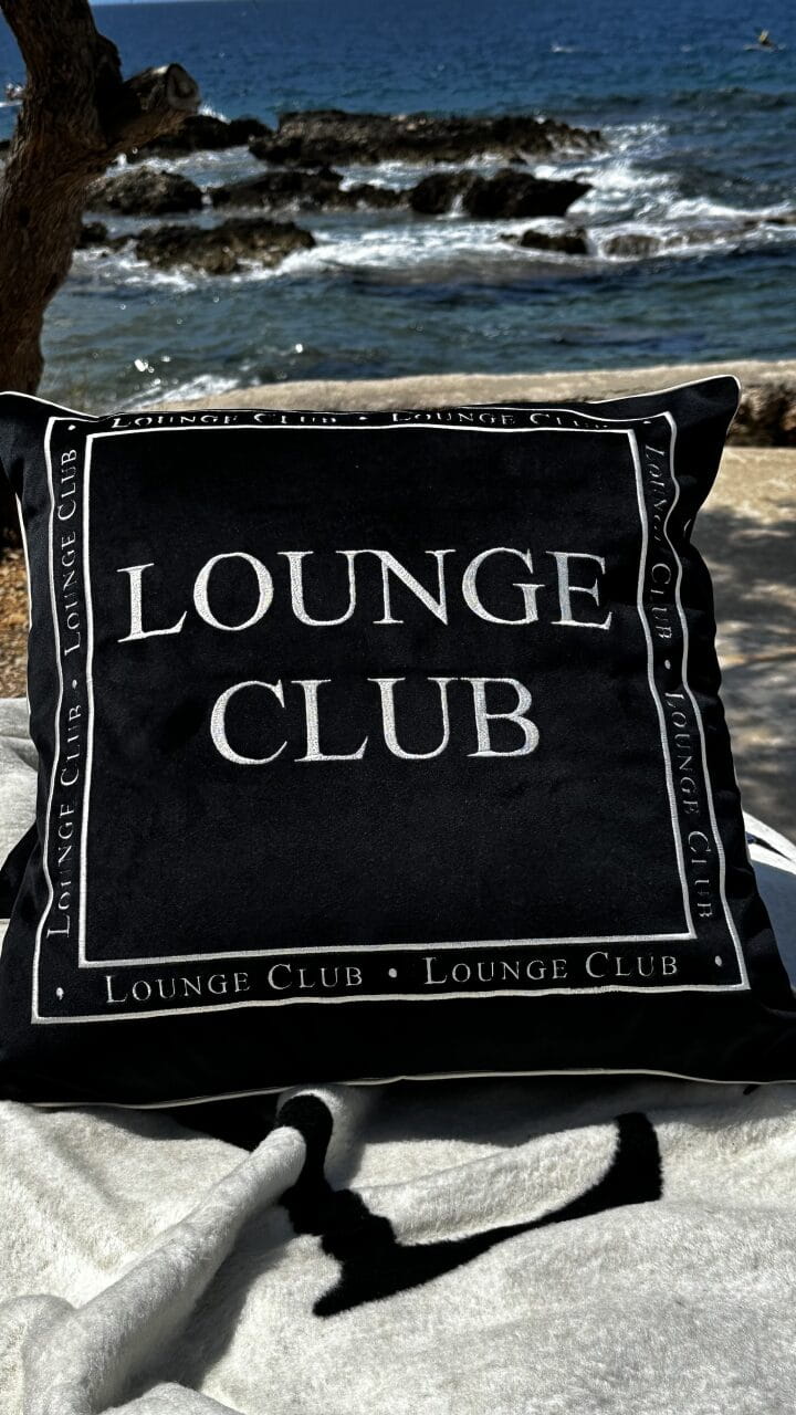 Poduszka haftowana Lounge Club w kolorze czarno-kremowym, z dużym napisem LOUNGE CLUB na froncie. Produkt leży na jasnym kocu, w tle widoczna jest woda i skały. Kształt kwadratowy, rozmiar standardowy, tkanina teksturowana.